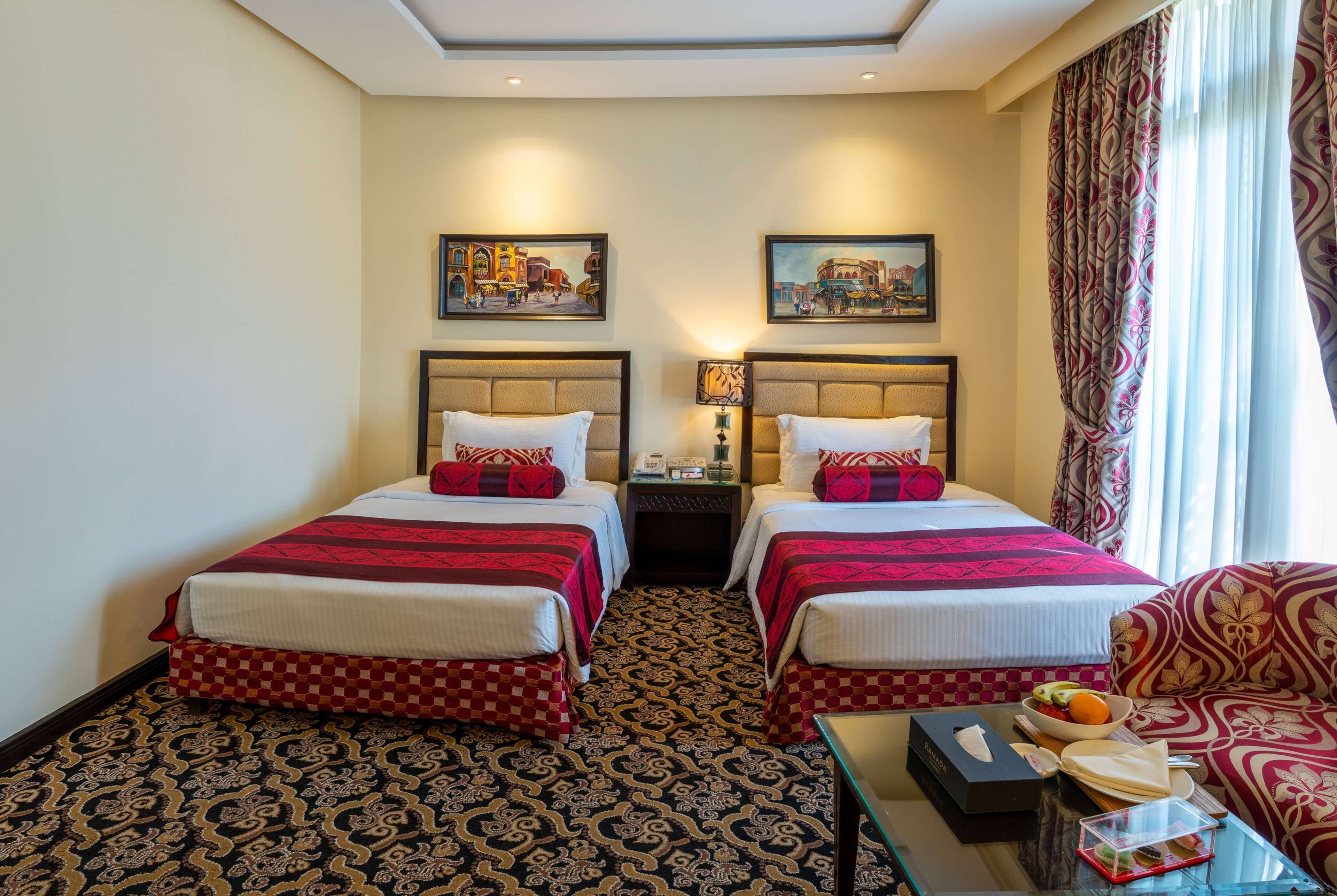 Hotelkamer met twee bedden, een geruit rode en witte sprei, een kamerbreed tapijt met dessin, een glazen bijzettafeltje met ontbijtartikelen, en twee schilderijen aan de muur.