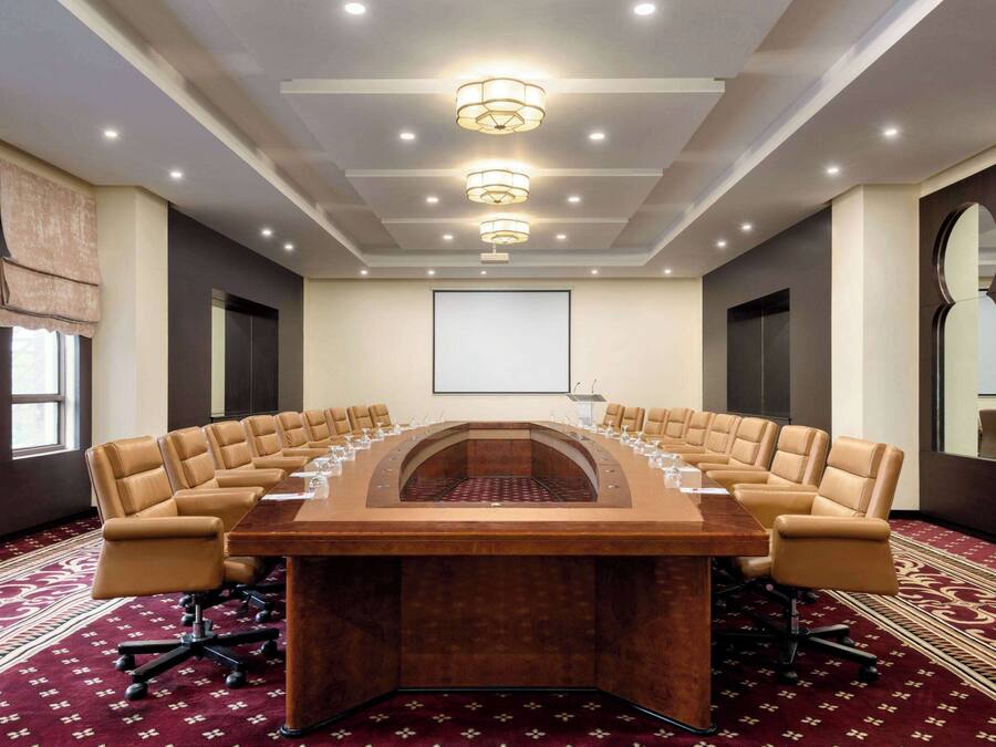 A conference room with a long table, multiple chairs, a projector screen, and a patterned carpet.