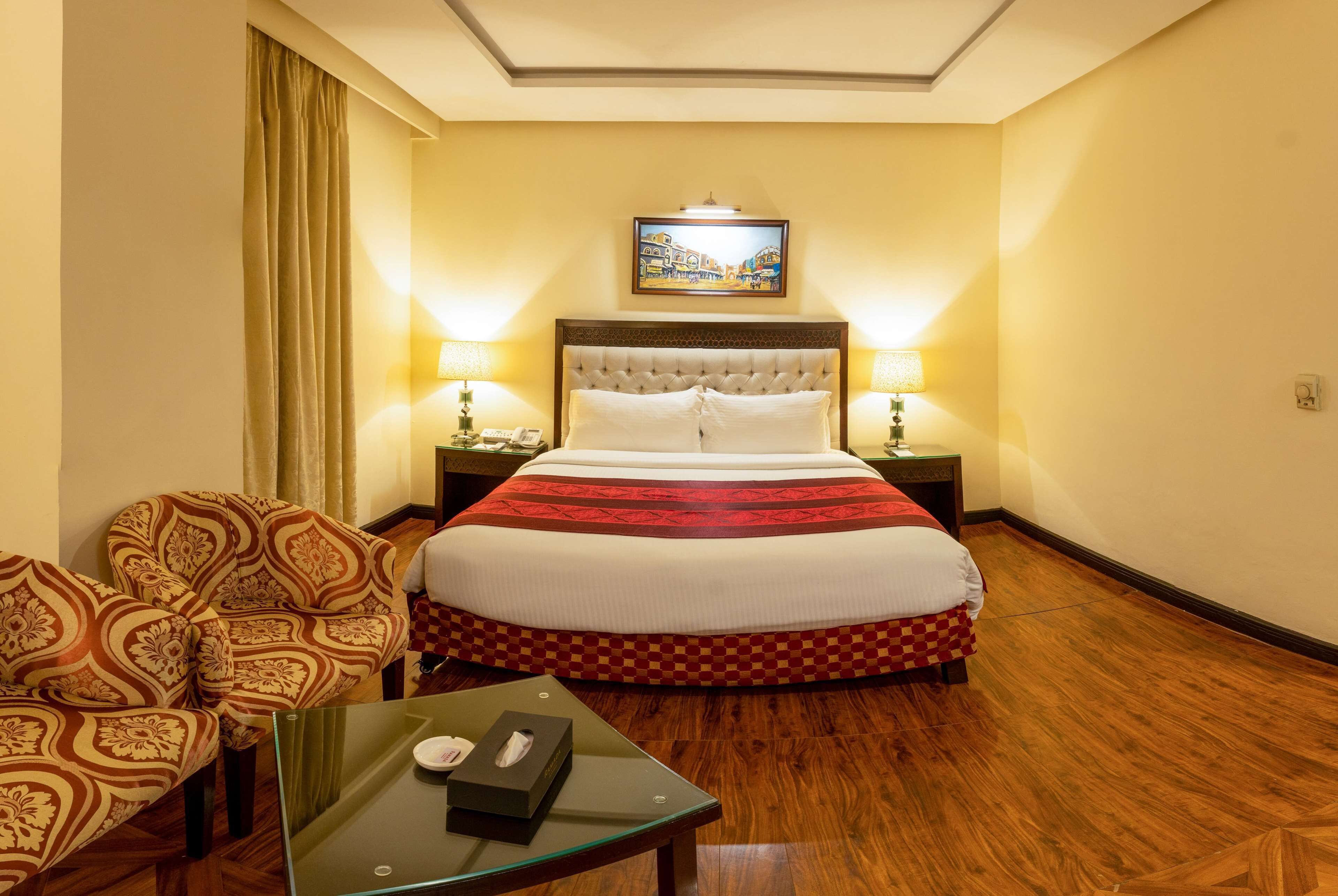 A hotel room with a large bed, two bedside tables with lamps, a glass coffee table, patterned armchairs, and a painting on the wall.