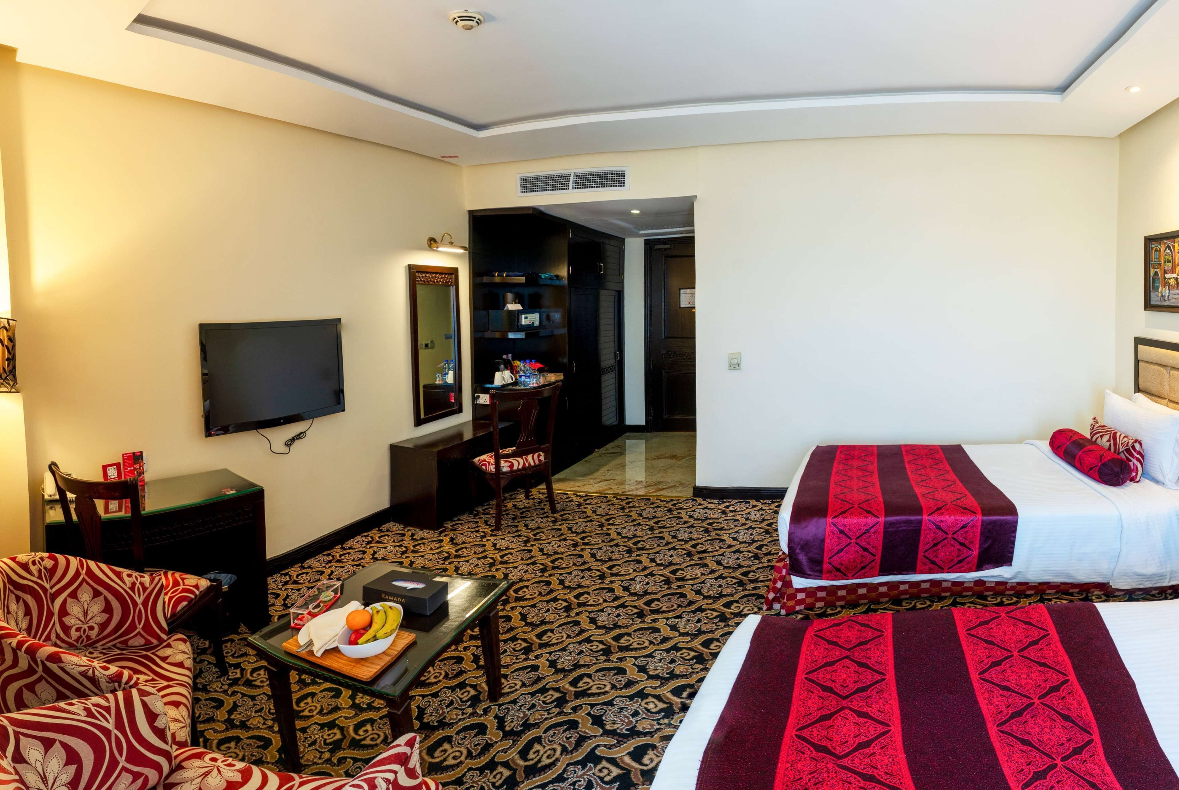 A hotel room with a TV, two beds with red and black bedding, a sofa, a small table with fruits, and a patterned carpet.