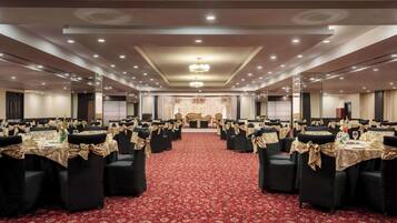 Uma sala de banquetes com mesas dispostas em fileiras, cada uma com cadeiras pretas e fitas douradas, e um chão coberto por um tapete vermelho.