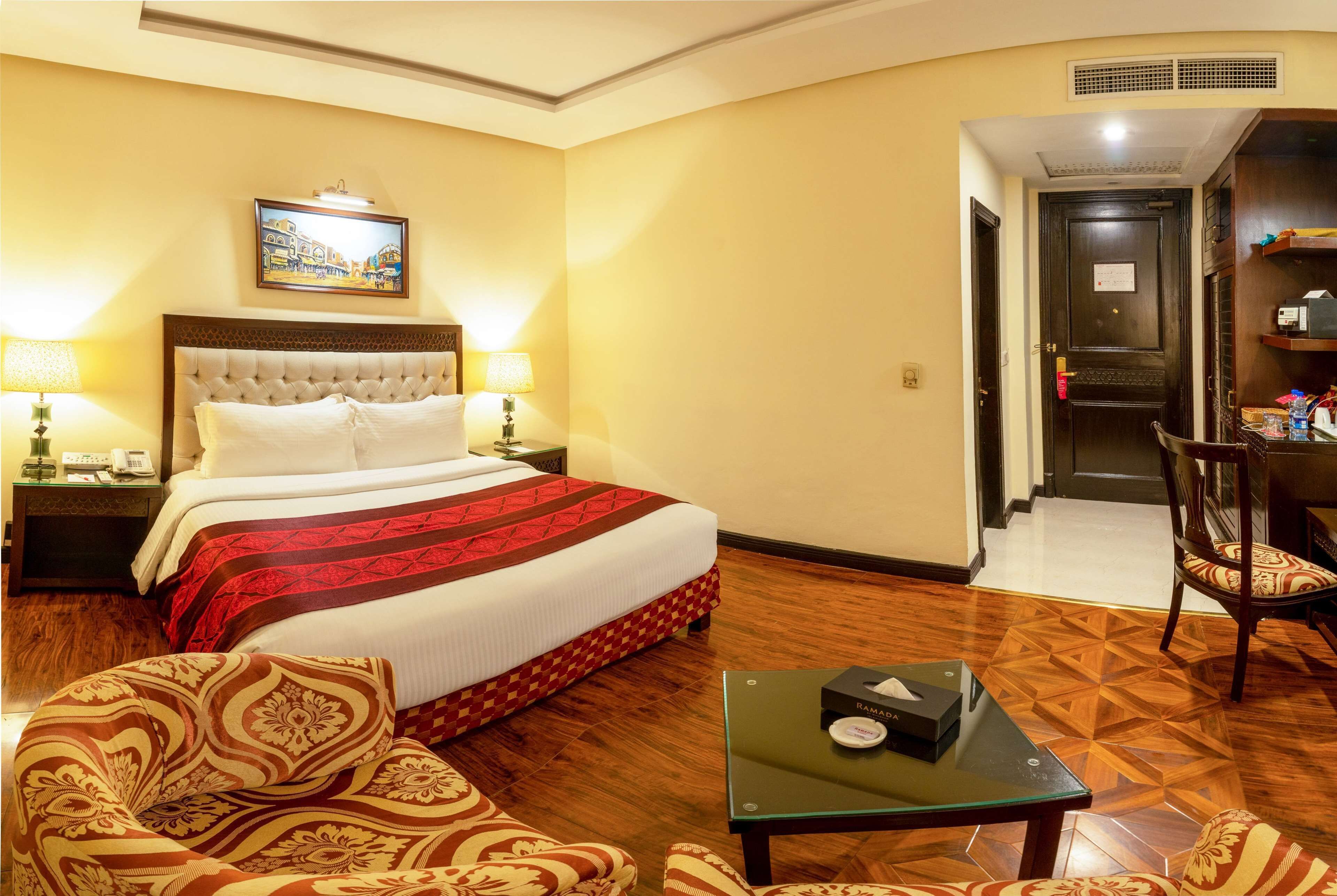 A hotel room with a large bed, a nightstand with a lamp, a chair, a table, and a door leading to a hallway.