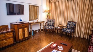 A hotel room with a wooden floor, a TV mounted on the wall, a desk with a lamp, two patterned armchairs, a small table, and a sofa. - Hotel Mehran (Karachi)