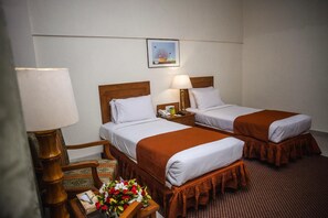 A hotel room with two single beds, a wooden chair, a nightstand with a lamp, a small table with a vase of flowers, and a framed picture on the wall.