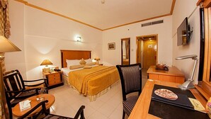 A hotel room with a bed, a desk, a chair, a lamp, a TV, and a door leading to another room. - Hotel Mehran (Karachi)