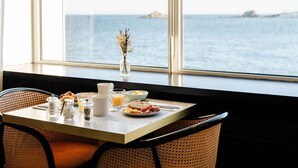 Une table de petit-déjeuner avec vue sur la mer, un vase rempli de fleurs séchées et des chaises en osier.