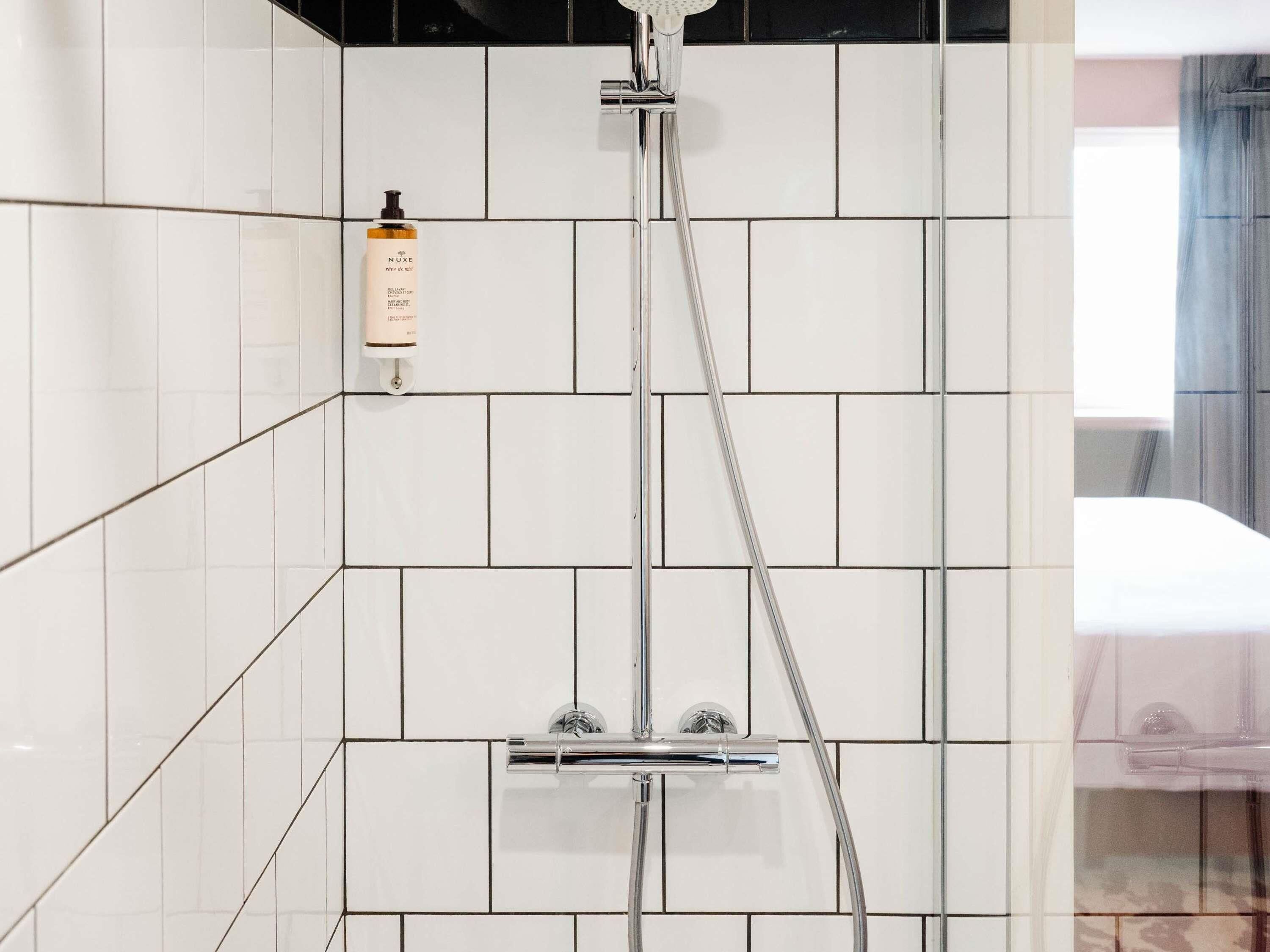 A modern bathroom with white tiles, a shower with a handheld showerhead, and a bottle of shampoo on the wall.