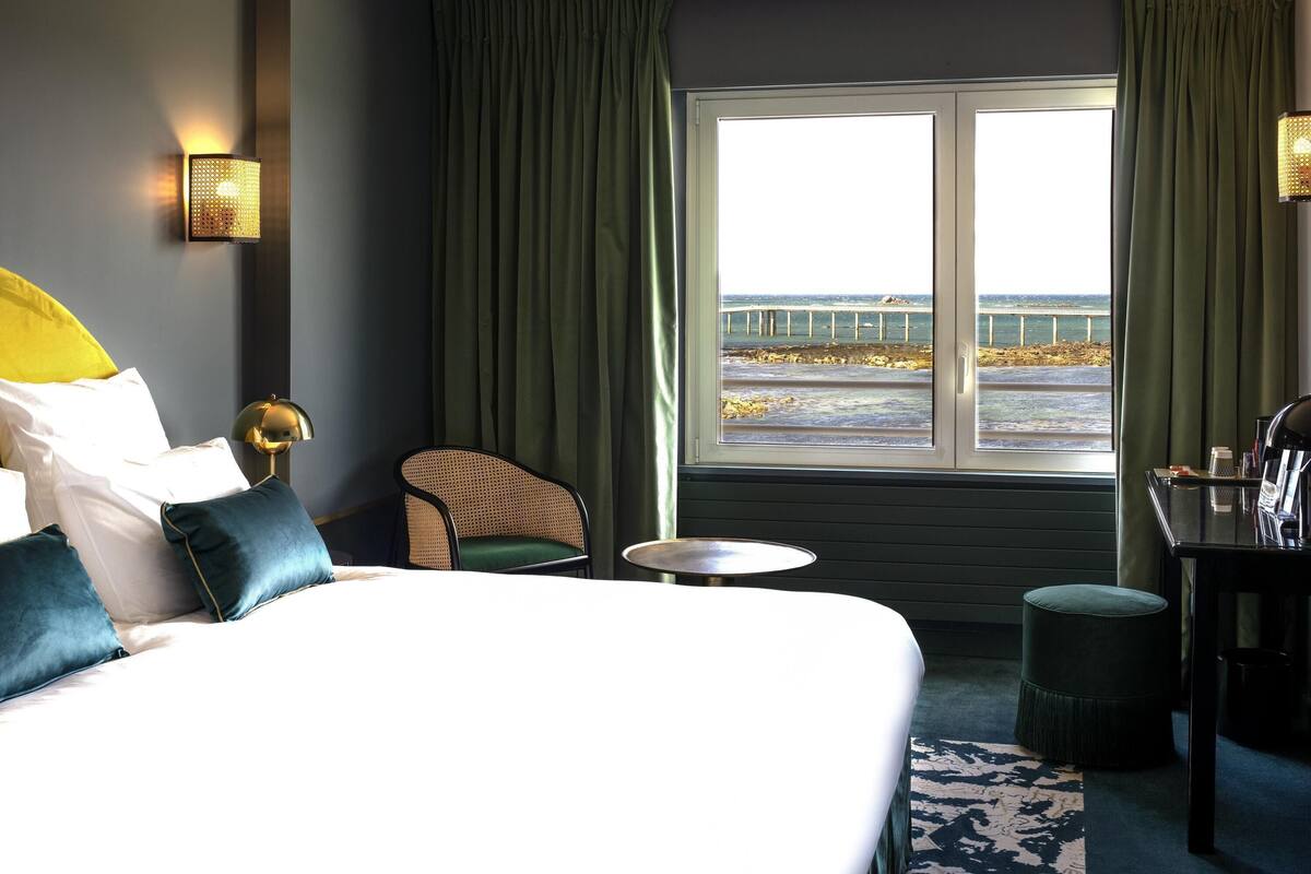 Een hotelkamer met een bed, een stoel, een tafeltje en uitzicht op zee door een raam.