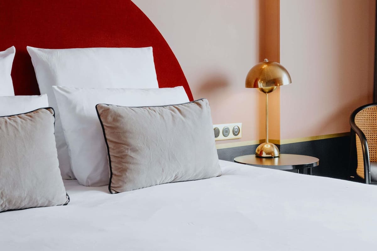 Een hotelkamer met een bed, kussens, een lamp, een stoel en een rode accentmuur.