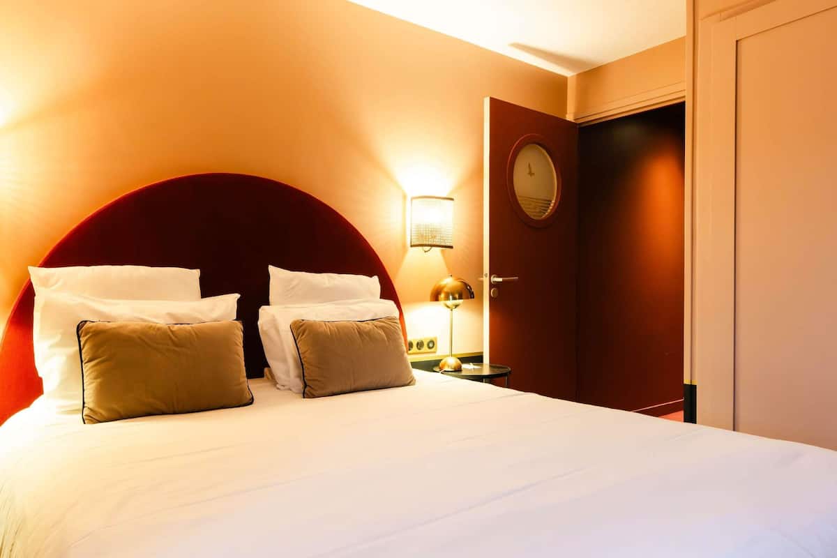 Een hotelkamer met een groot bed, een ronde spiegel, een nachtkastje met een lamp en een deur met een ronde spiegel.