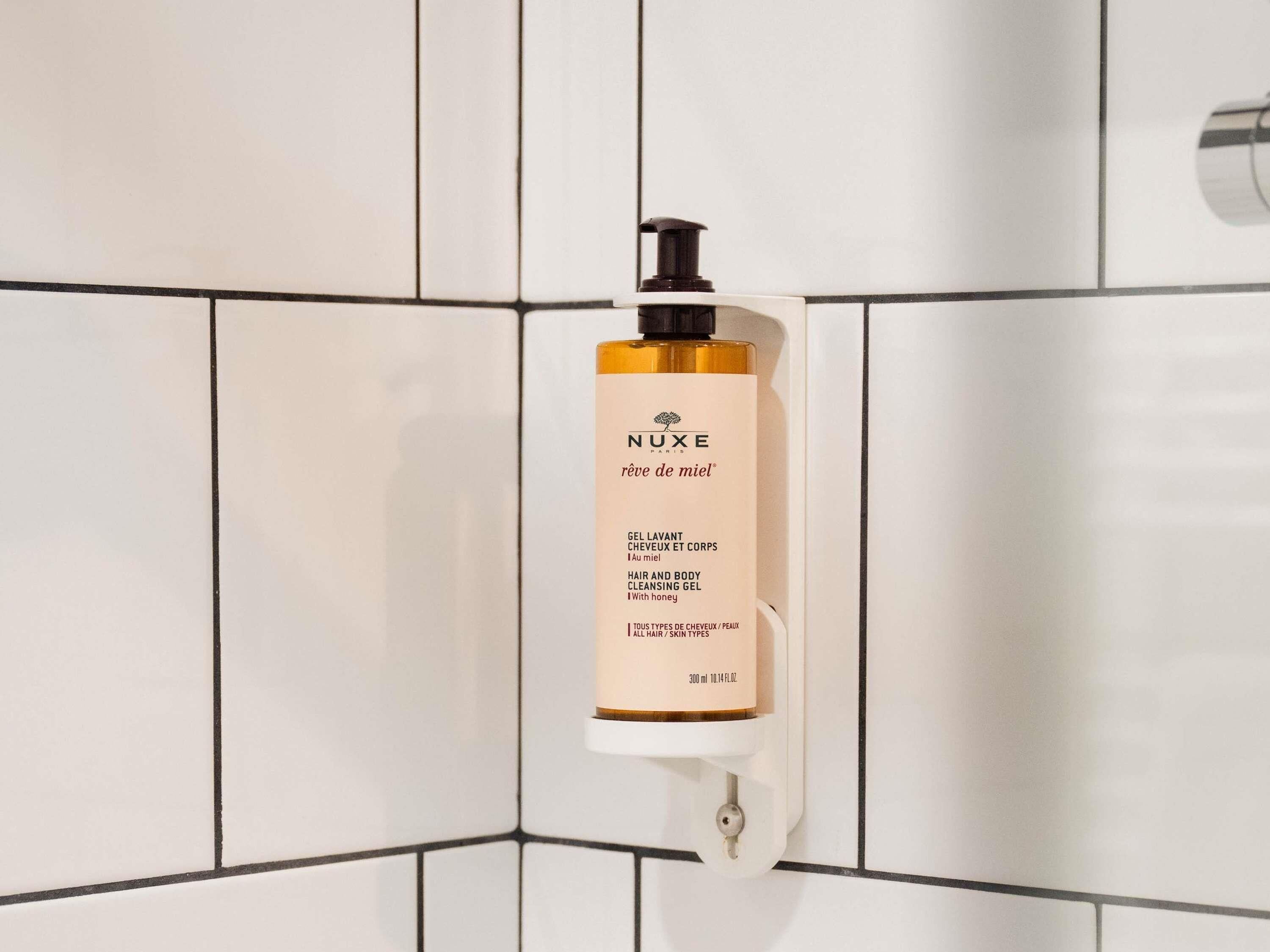 A bathroom with white tiled walls and a white shelf holding a bottle of NUXE Paris hair and body cleansing gel.