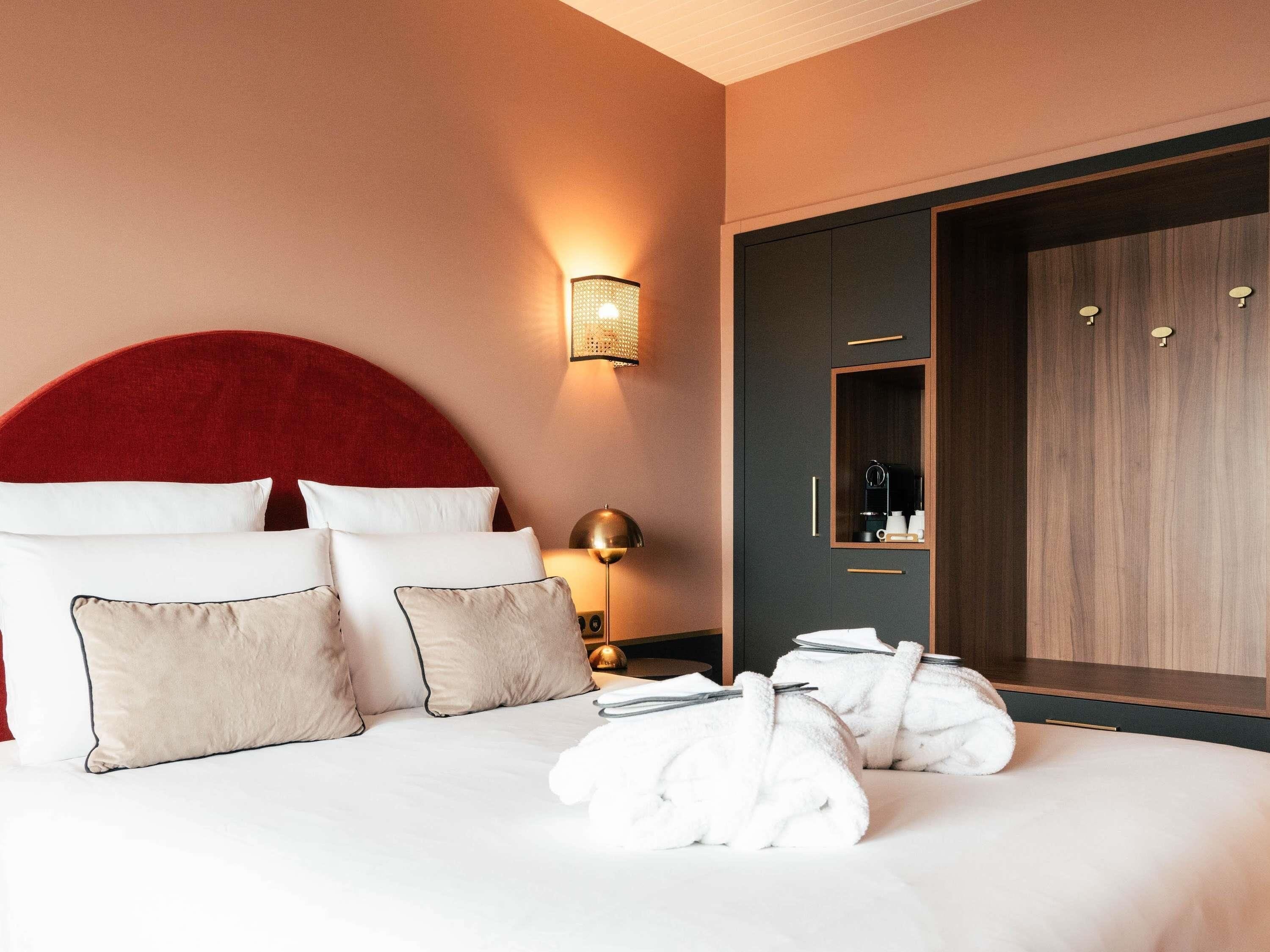 A hotel room with a large bed, multiple pillows, a red headboard, a bedside lamp, a wardrobe, and a coffee machine.