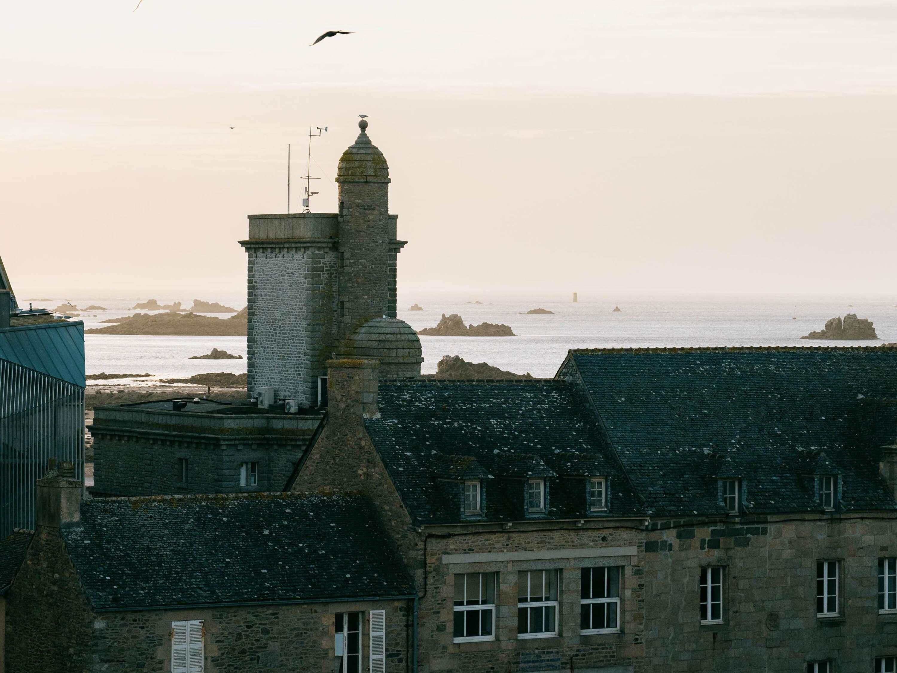 Eine Küstenstadt mit Steinbauten, einem Leuchtturm und felsigen Landspitzen im Hintergrund.