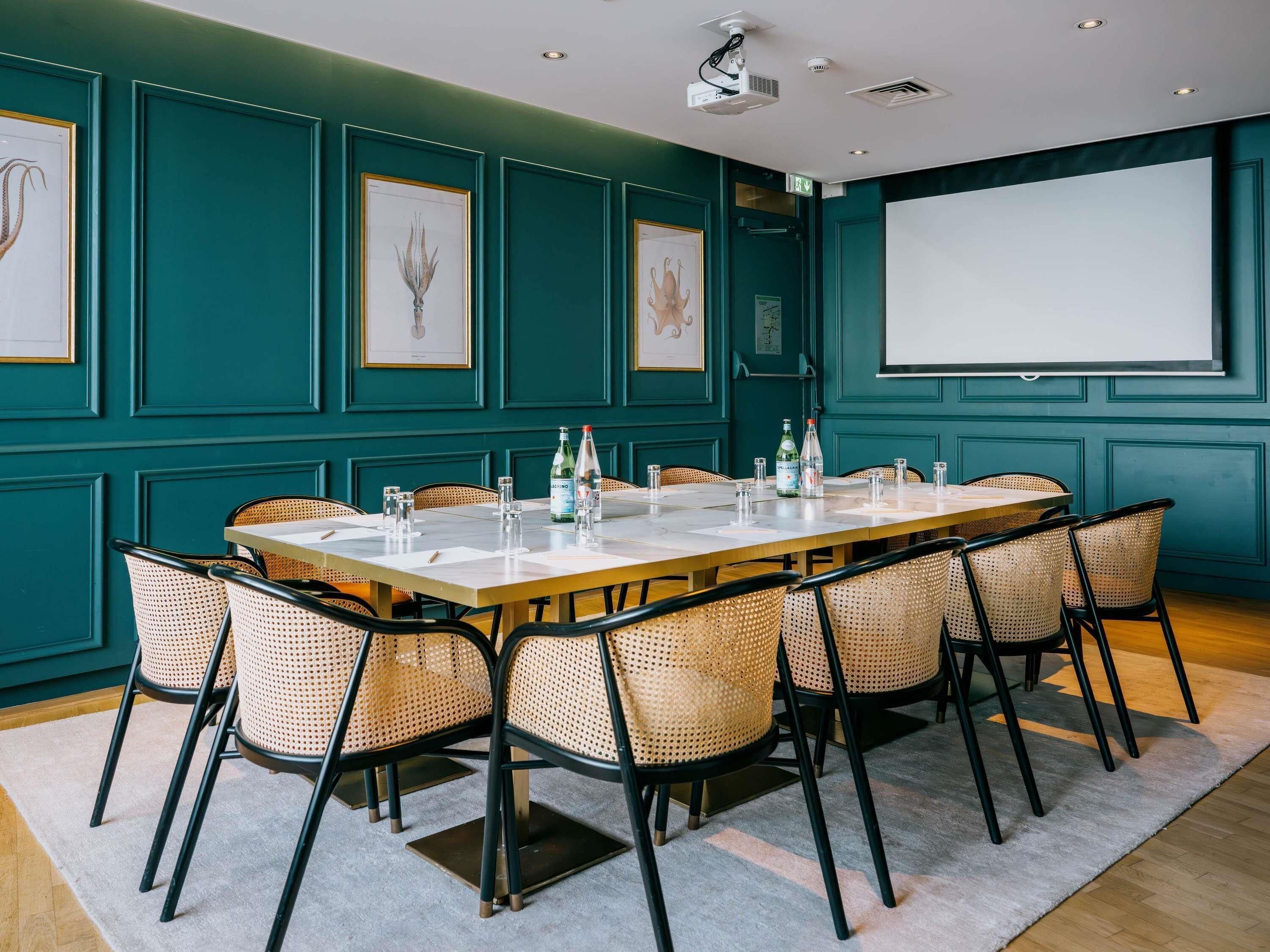 Une salle de conférence avec une longue table, des chaises en osier, un écran de projection et des reproductions botaniques encadrées sur des murs de couleur turquoise.