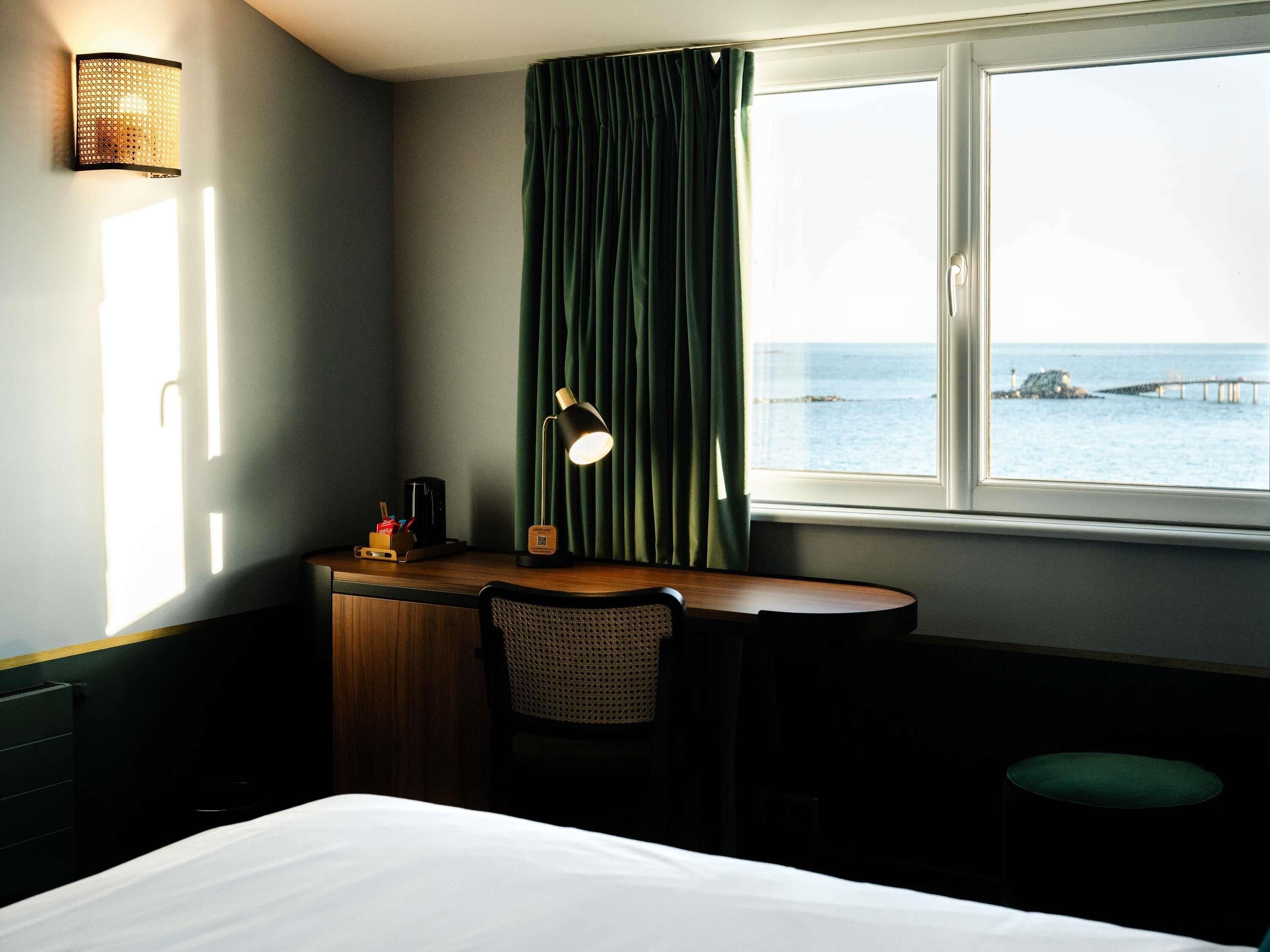 A hotel room with a bed, a desk with a lamp, a chair, a window with curtains, and a view of the sea.