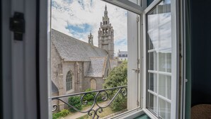 Une vue depuis une fenêtre donnant sur un balcon en fer forgé, avec en contrebas un bâtiment historique doté d’une tour imposante et une église surmontée d’un toit en tuiles.