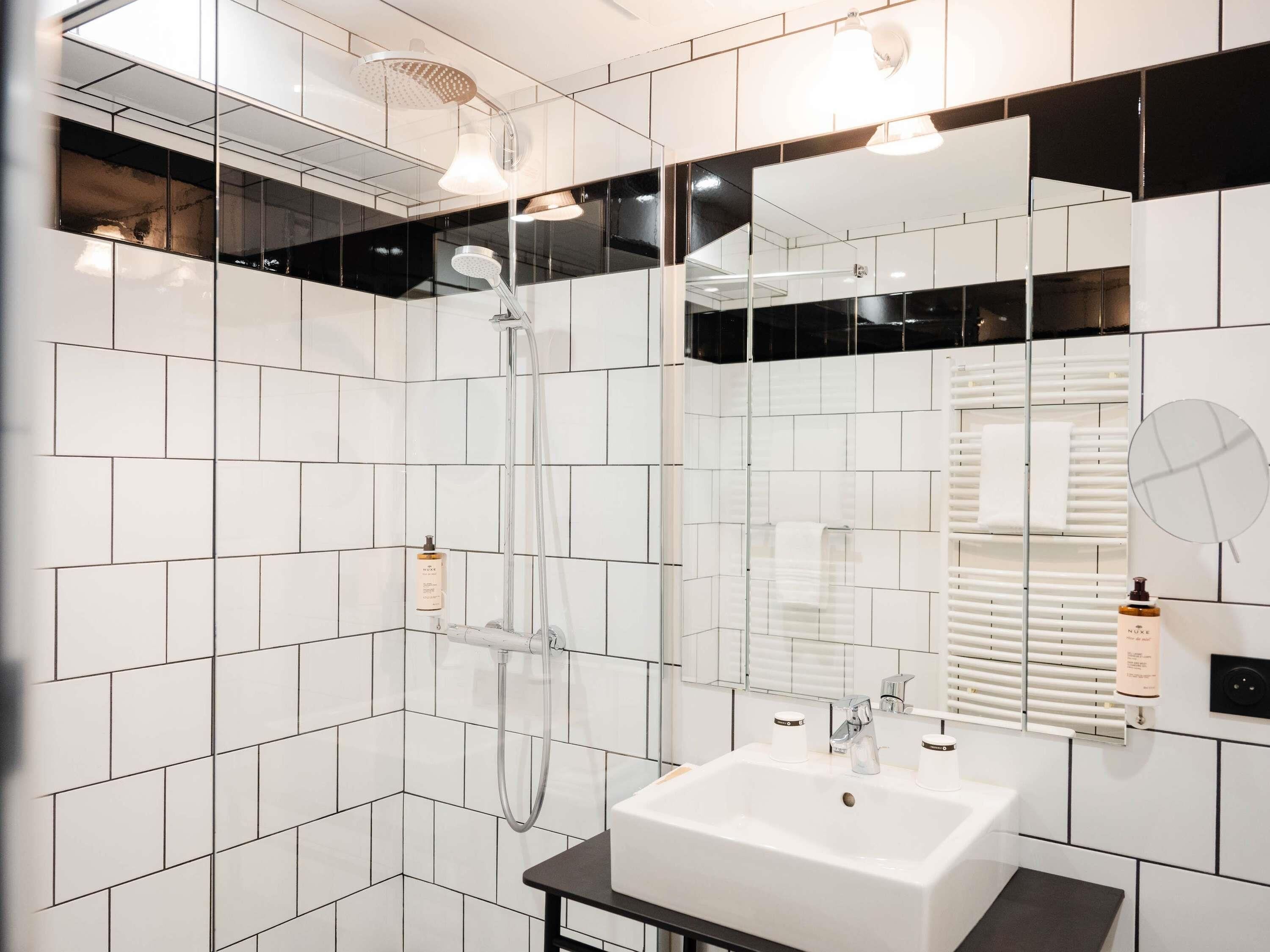 Un baño moderno con azulejos blancos, ducha con mampara de vidrio, lavamanos blanco y un espejo.