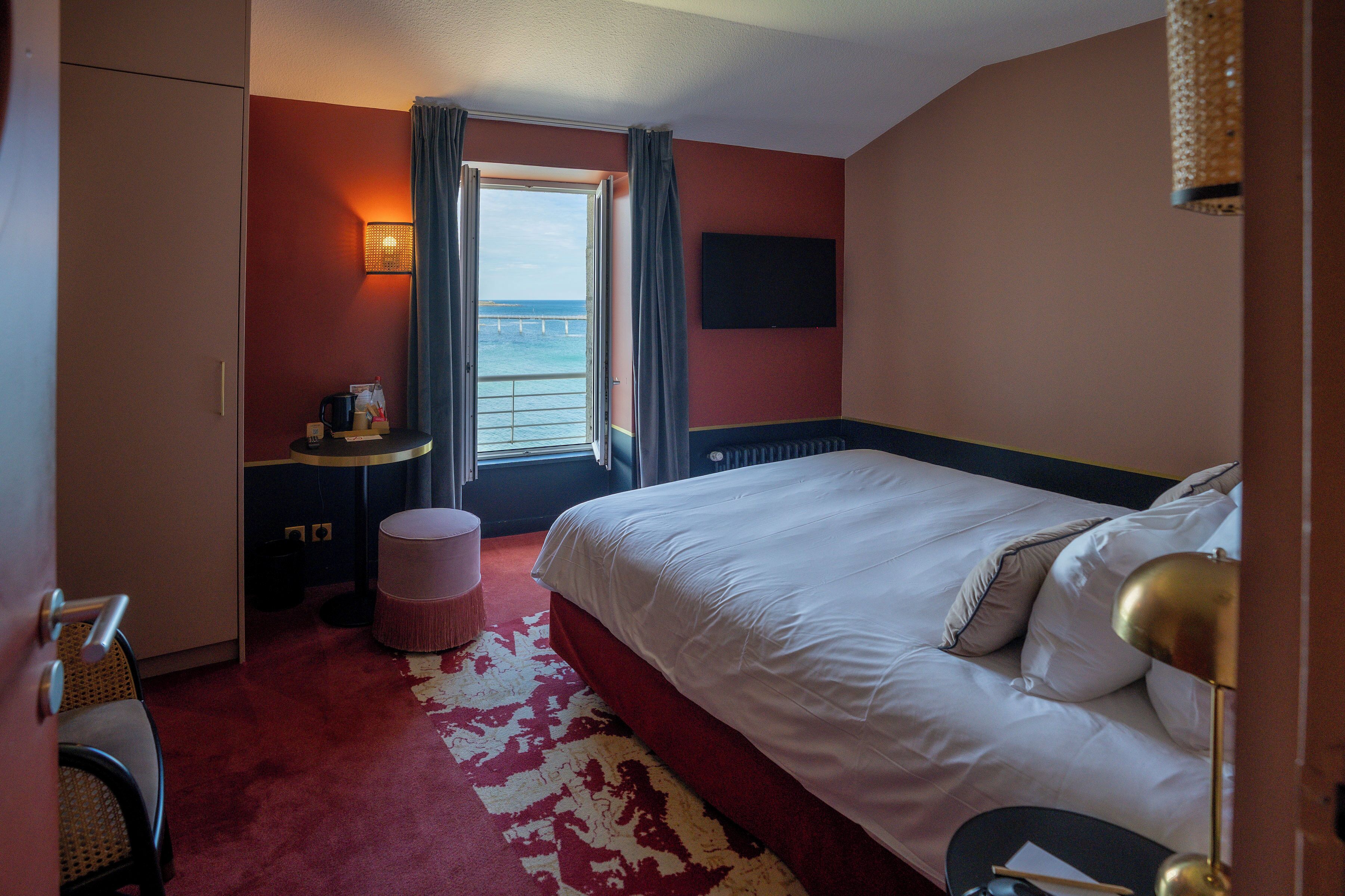 A hotel room with a bed, a desk, a chair, a small stool, a television, and a view of the sea through a window.