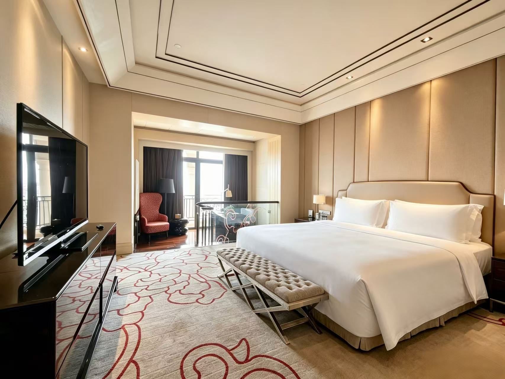 Quarto de hotel com uma cama grande, televisão, área de estar com uma poltrona e uma mesa de vidro.