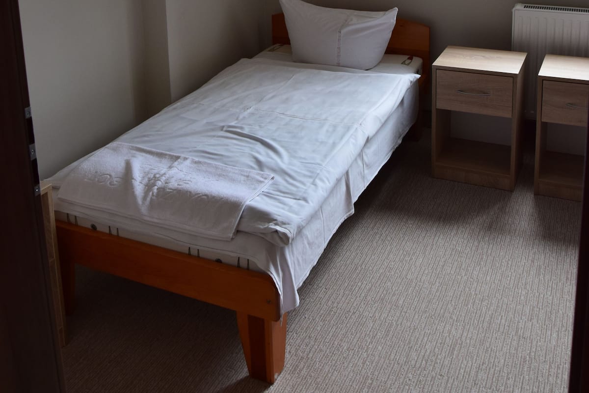 Une petite chambre avec un lit simple, un cadre en bois, une table de chevet avec des tiroirs, un radiateur et une fenêtre donnant sur un paysage verdoyant.