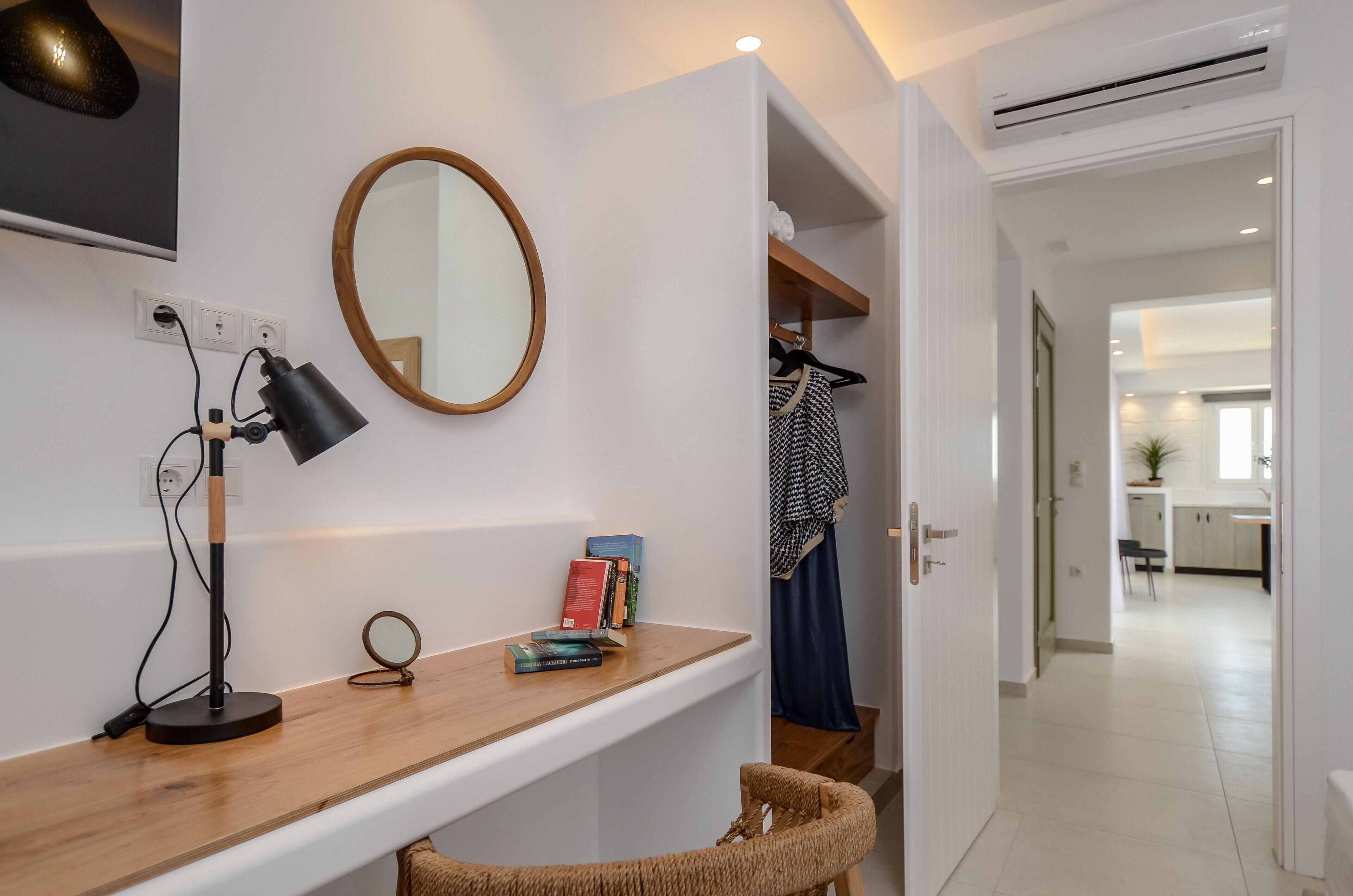A modern room with a wooden desk, a round mirror, a hanging coat, and a view into a kitchen area.