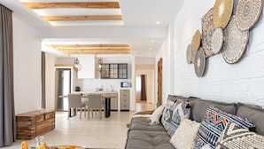 Una sala moderna con un sofá gris, cojines estampados y una mesa de centro de madera con fruta y bebidas. Se ve una cocina al fondo, junto con una zona de comedor.