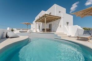 Un espace moderne aménagé au bord de la piscine, avec une piscine à l’eau d’un bleu limpide, une architecture blanche et des sièges d’extérieur.
