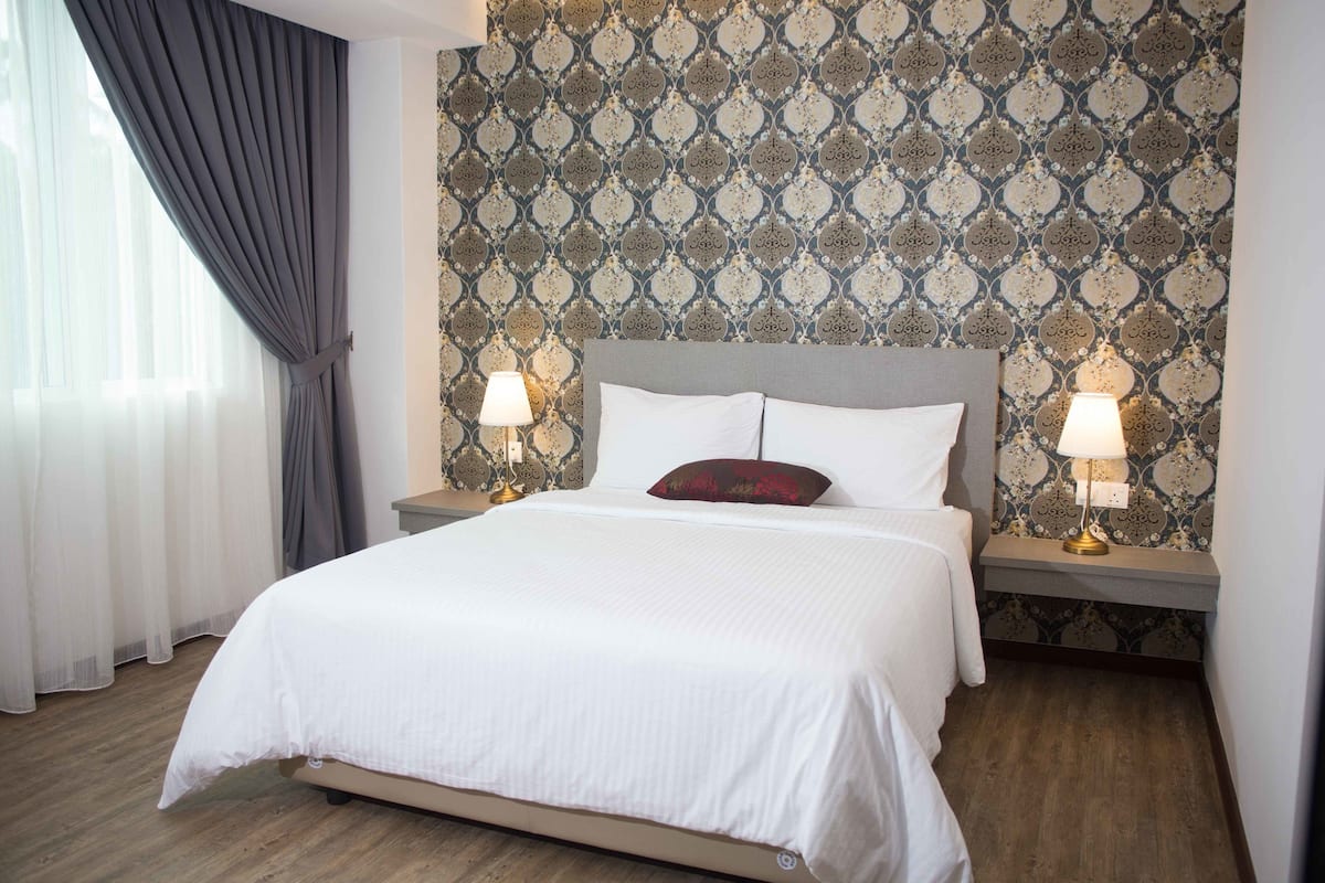 Cama bem arrumada com roupa de cama branca, duas luminárias de cabeceira e um papel de parede estampado.