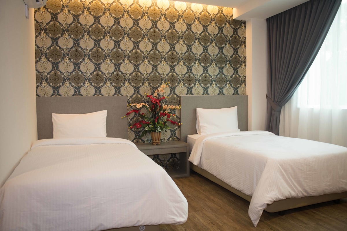 Quarto de hotel com duas camas, mesa de cabeceira com um vaso de flores, papel de parede estampado e janela com cortinas.