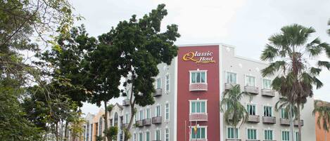 Un hotel de varios pisos con balcones, un cartel llamativo que dice "Classic Hotel", y una vista de la calle con coches aparcados y árboles.