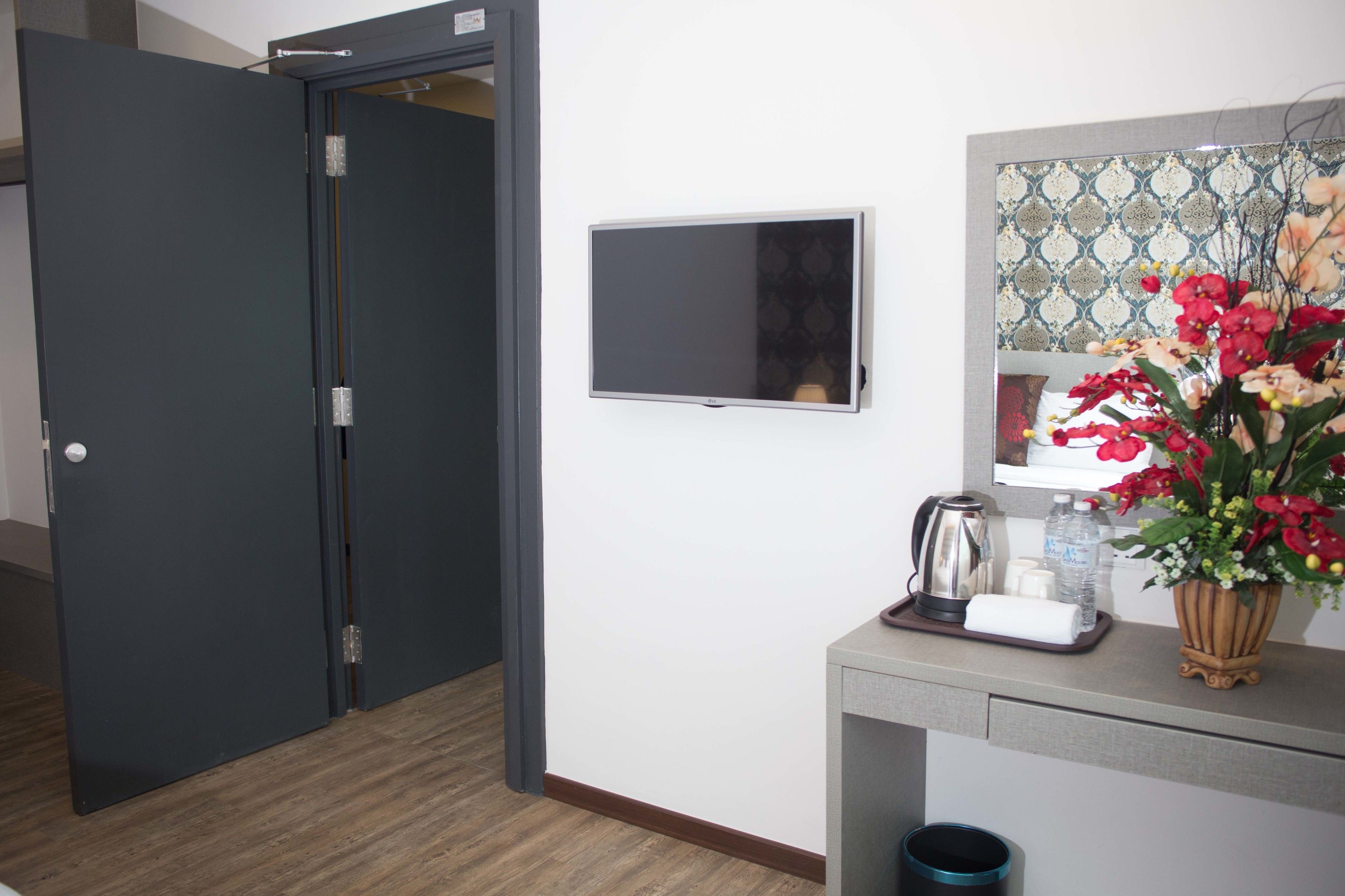 A hotel room with a closed door, a flat-screen TV mounted on the wall, a small table with a kettle, a vase with flowers, and a trash bin.