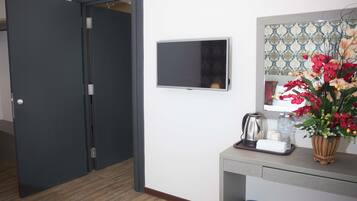 A hotel room with a closed door, a flat-screen TV mounted on the wall, a small table with a kettle, a vase with flowers, and a trash bin.