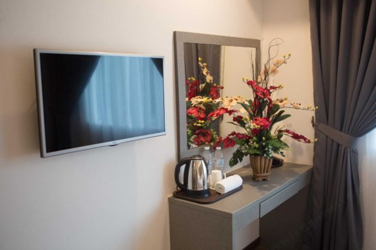 A hotel room with a flat-screen TV mounted on the wall, a mirror above a vanity with a floral arrangement, and a coffee maker on the counter.