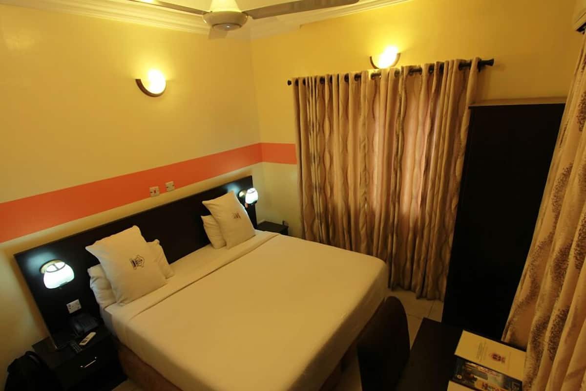 Una habitación de hotel con cama, lámparas de noche, mesita de noche, maleta y cortinas.