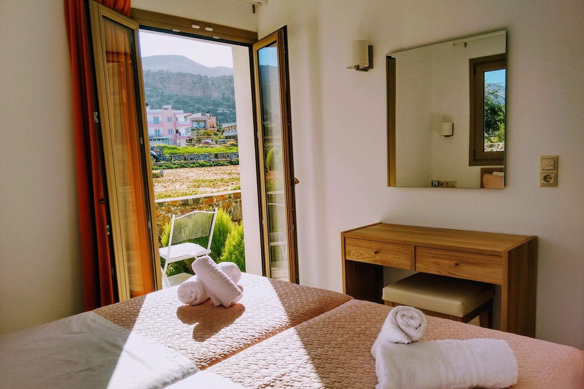 A hotel room with a bed, a wooden dresser, a mirror, and a view of buildings and greenery outside.