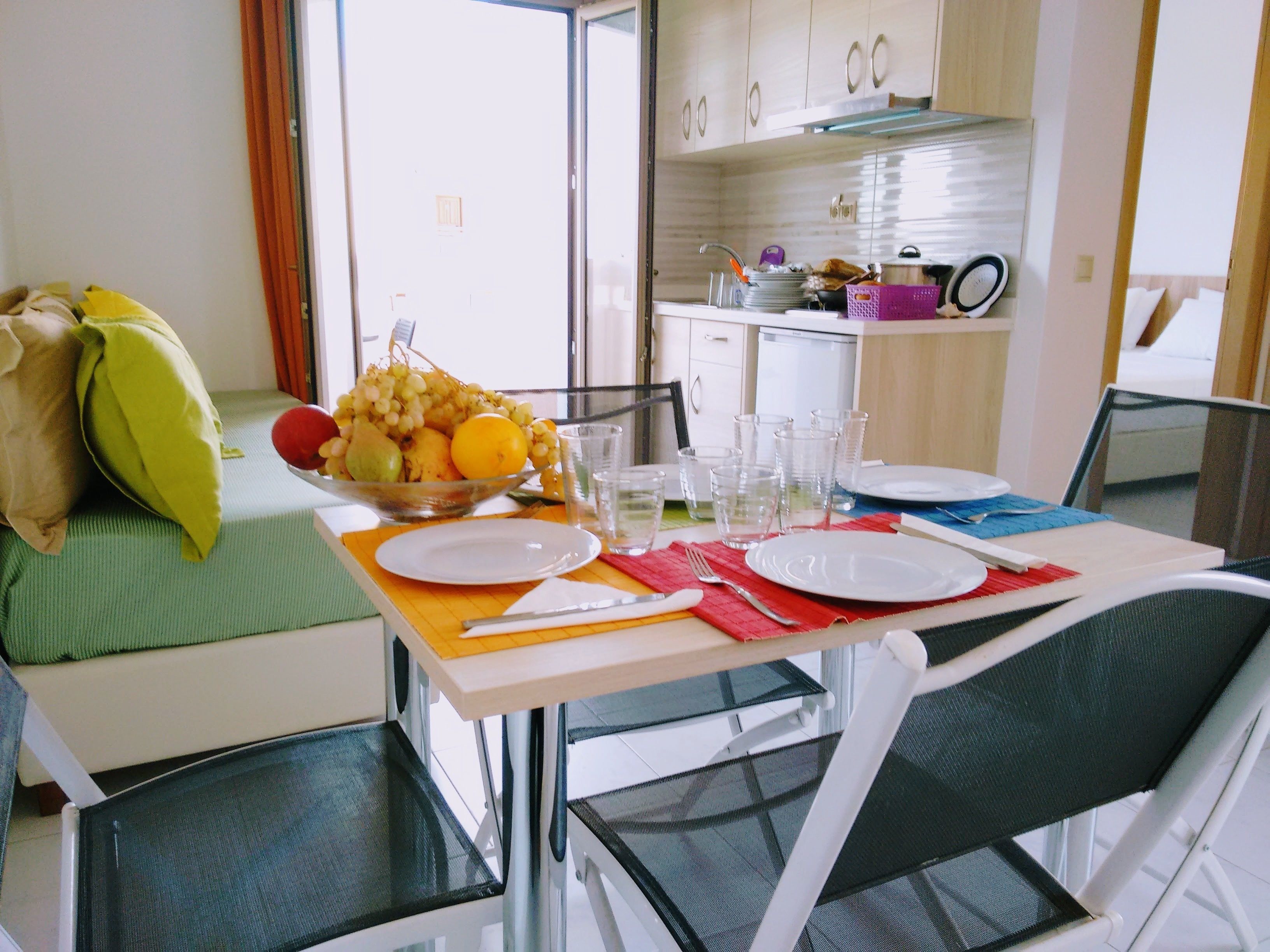 Un coin repas avec une table dressée pour deux personnes, un saladier de fruits et une cuisine en arrière-plan.
