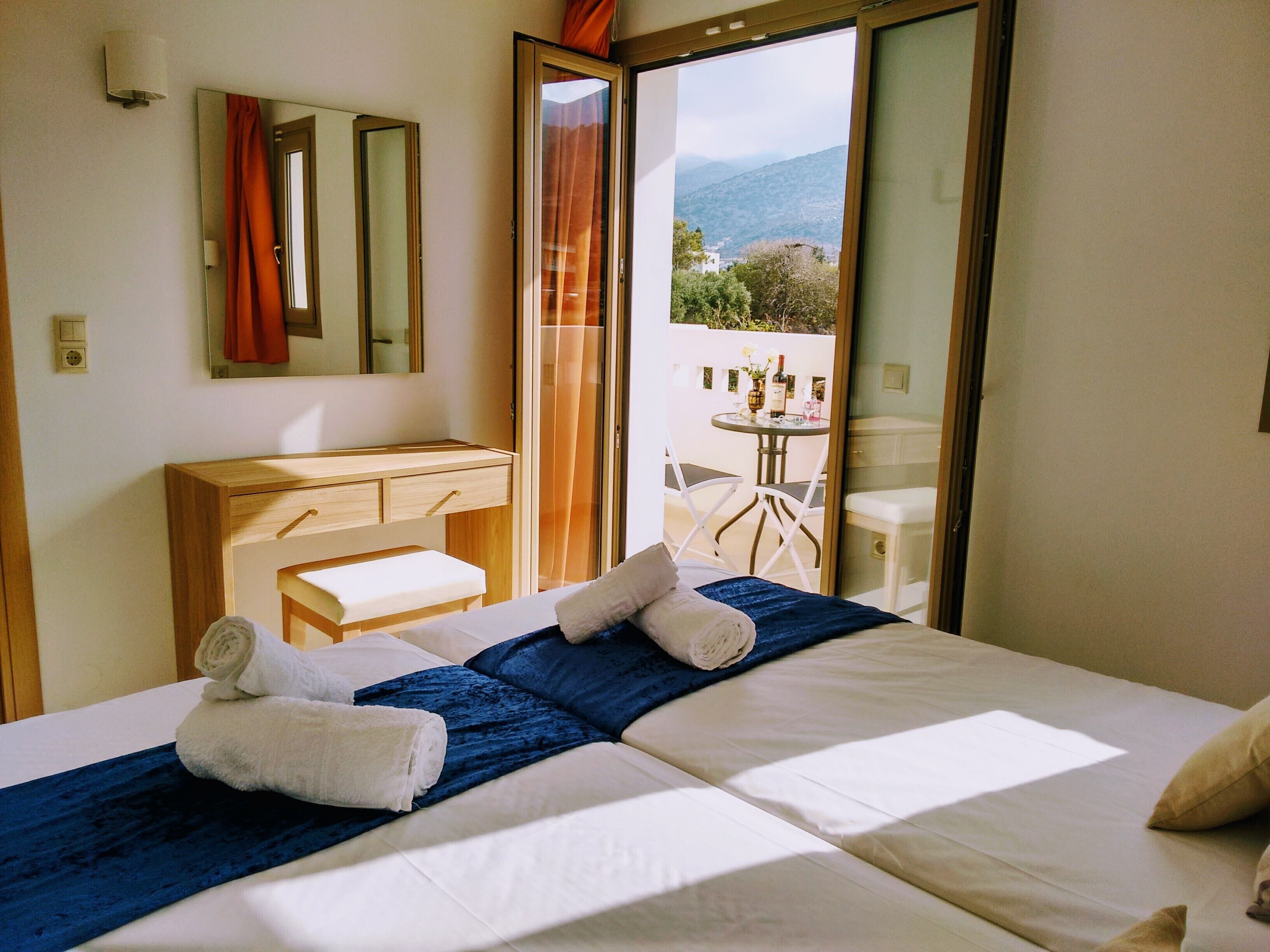 Habitación de hotel con cama, cómoda de madera, espejo y balcón con mesa y sillas.
