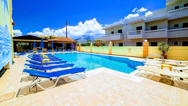 A swimming pool with blue lounge chairs, a covered seating area, and a building with balconies. - Casa Malena (Hersonissos)