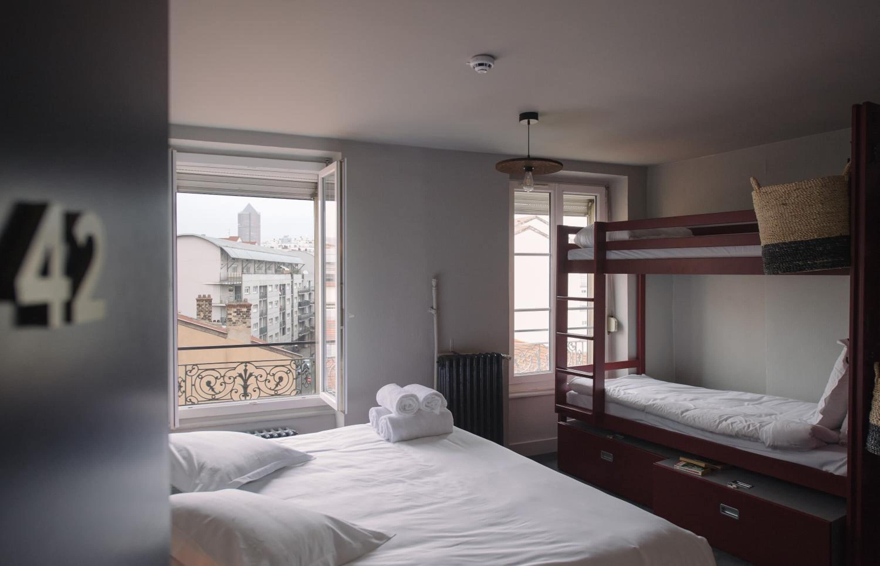 Une chambre d’hôtel avec deux lits, une vue sur les immeubles à travers de grandes fenêtres et un balcon avec une balustrade.