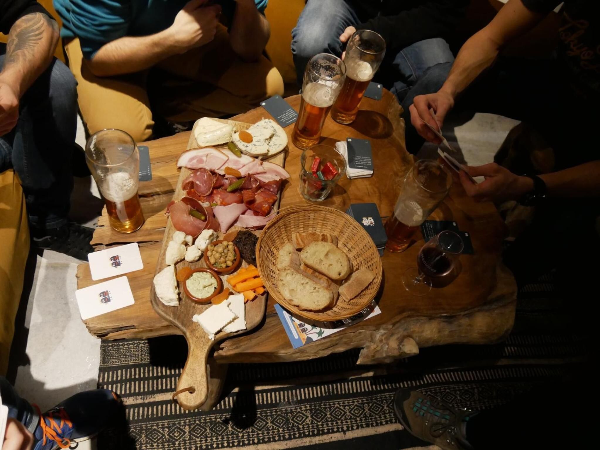 A wooden table with food and drinks, including cheese, meats, and bread, in a casual dining setting.