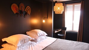 Une chambre d’hôtel avec un lit, des oreillers blancs, un plaid à motifs, une lampe fixée au mur et une fenêtre avec des rideaux.