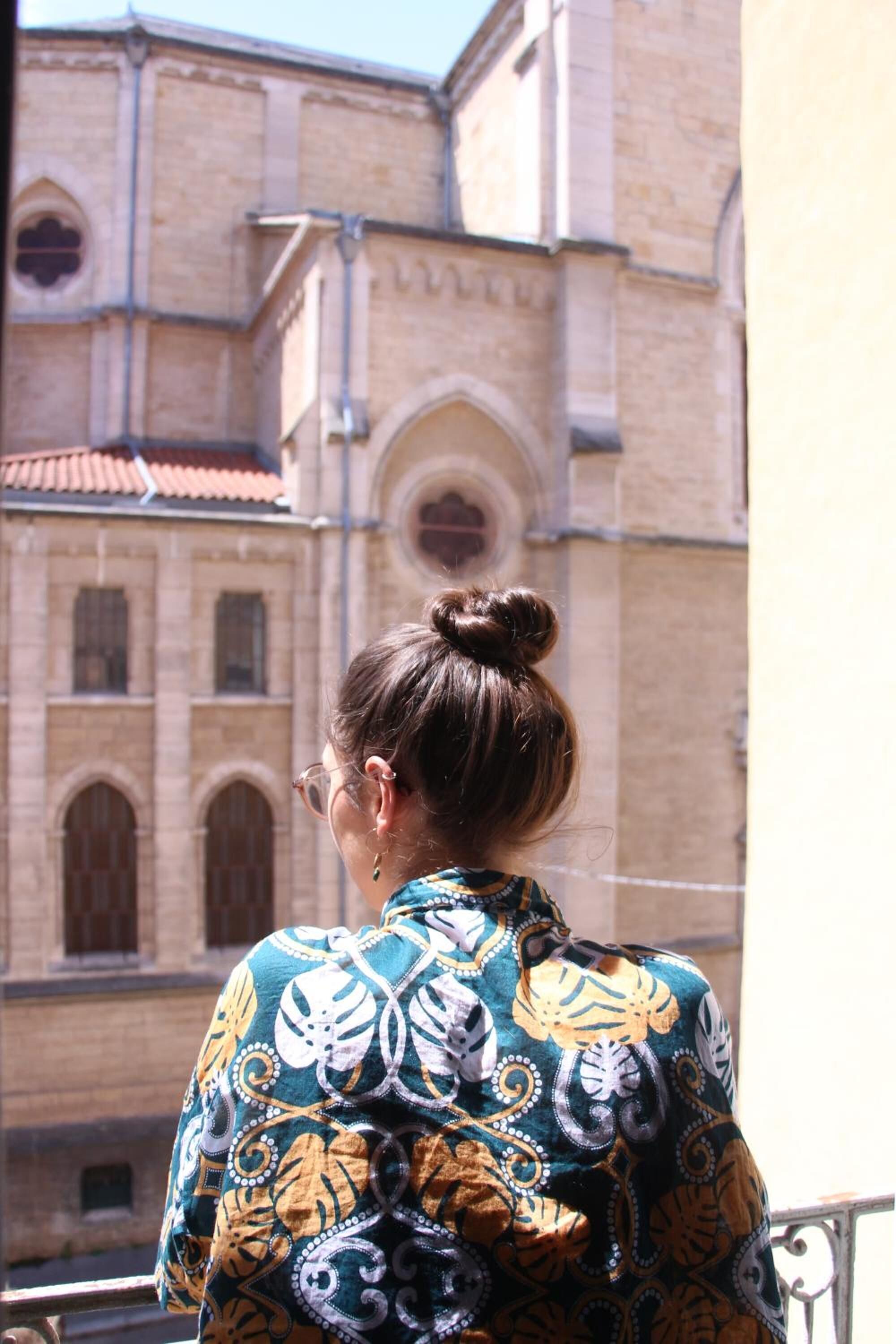 Una persona de pie en un balcón, con una camisa estampada, mirando un edificio con ventanas arqueadas y tejado de tejas.
