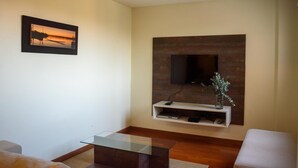 A modern living room with a flat-screen TV mounted on a wooden wall, a glass coffee table, and a framed picture on the wall. - Andares Del Agua (Villa Elisa)