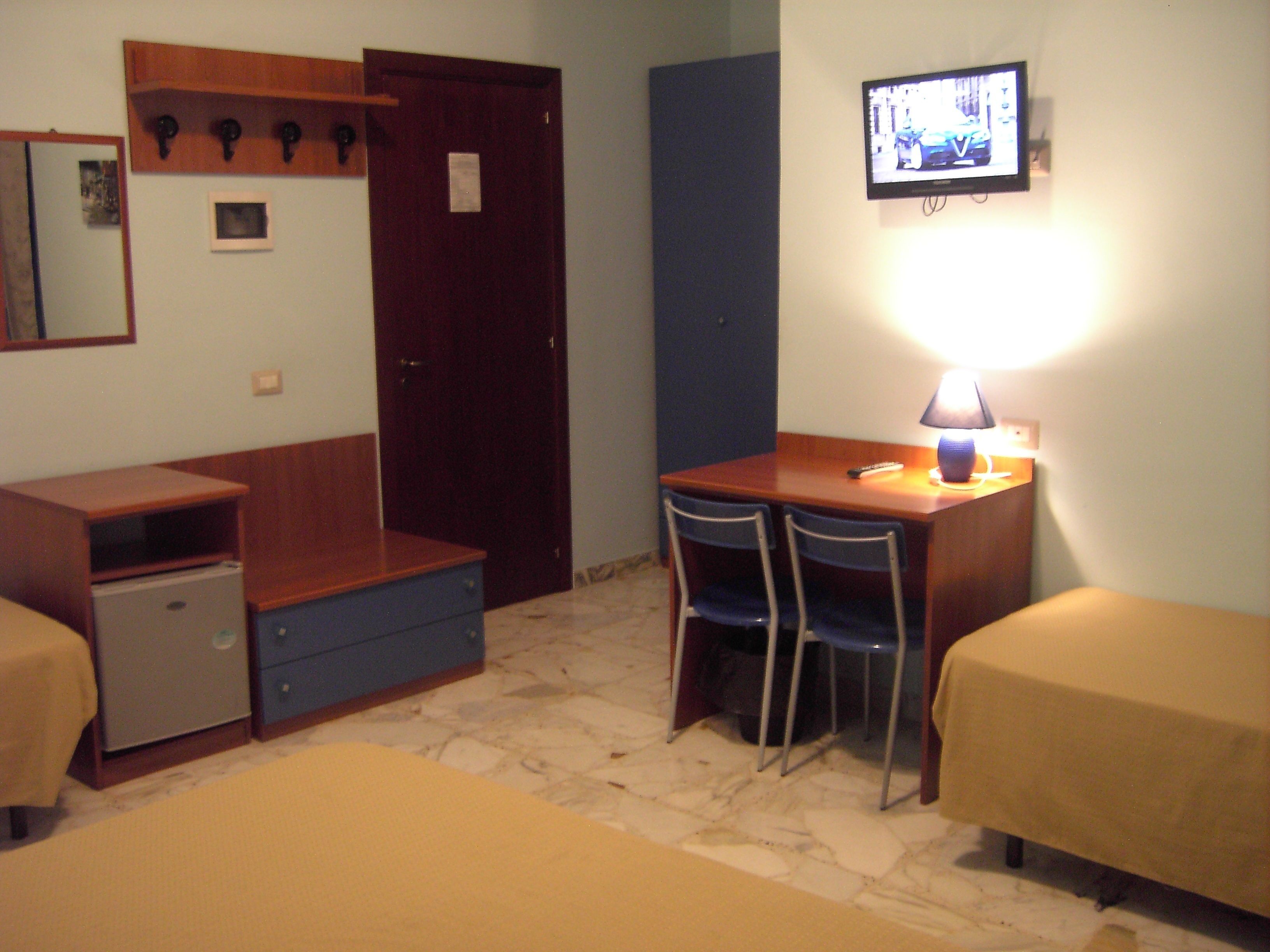 A small, compact room with a desk, two chairs, a bed, a wardrobe, and a television.