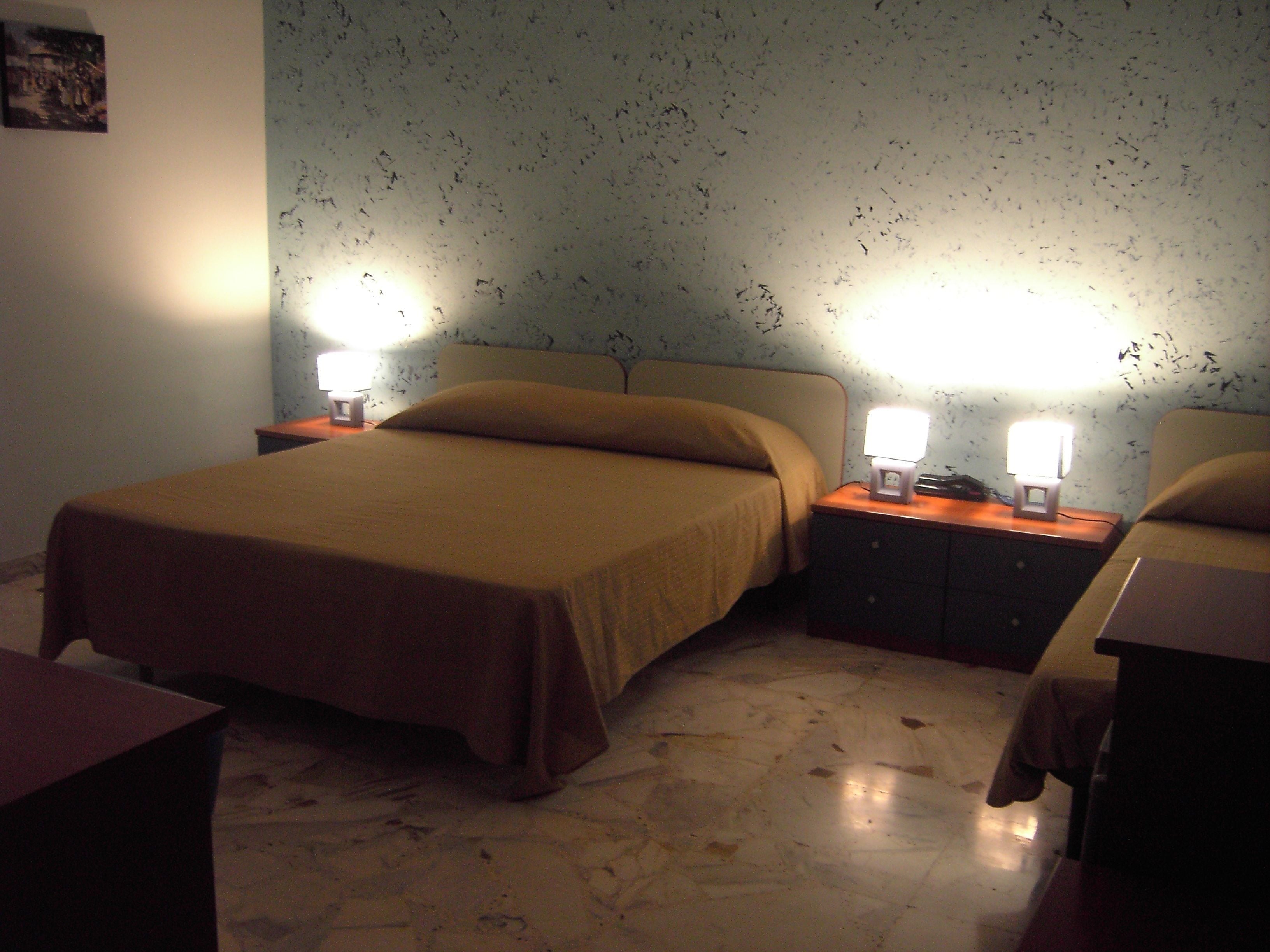 A bedroom with a bed, two bedside tables with lamps, a nightstand, and a wall with a pattern.