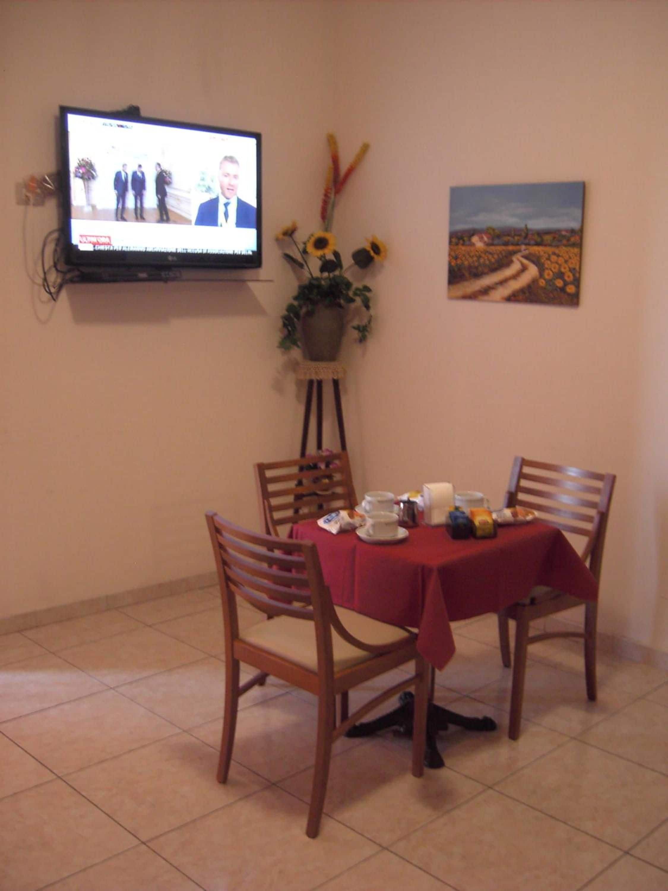 Ein Essbereich mit an der Wand montiertem Fernseher, der eine Nachrichtensendung zeigt, einem Tisch, der mit einer roten Tischdecke gedeckt ist, und einem Gemälde einer Sonnenblumenwiese an der Wand.