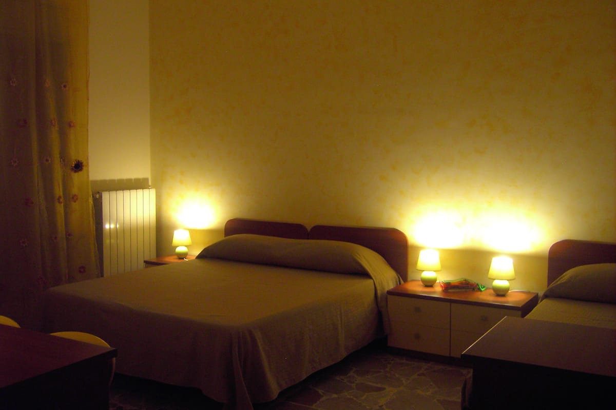 A hotel room with two beds, a nightstand with lamps, a curtain, and a radiator.