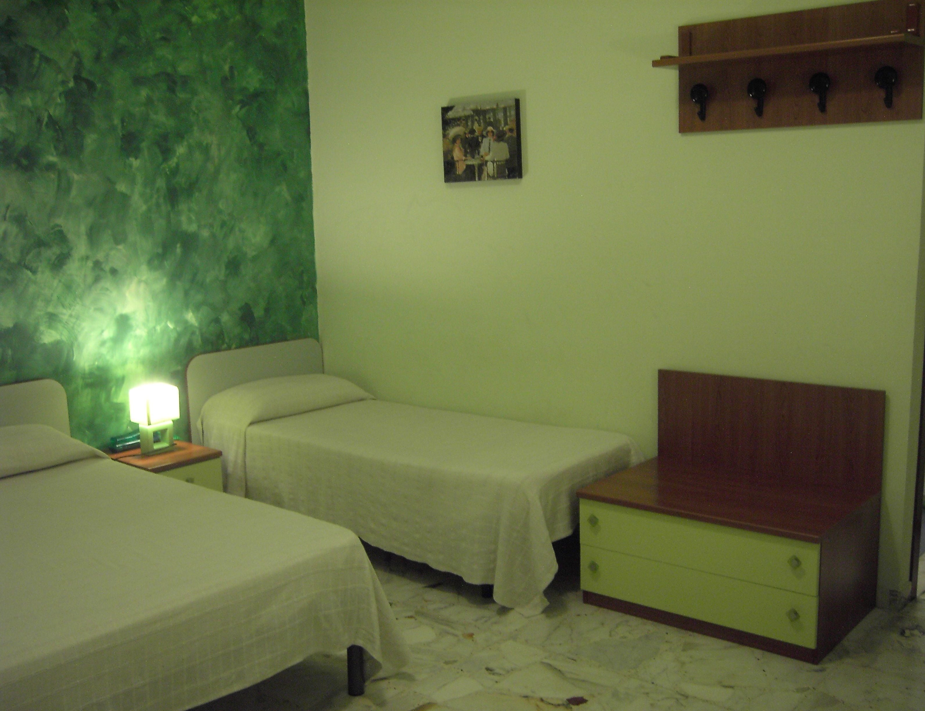 A room with two single beds, a bedside table with a lamp, a small dresser, and a green textured wall.