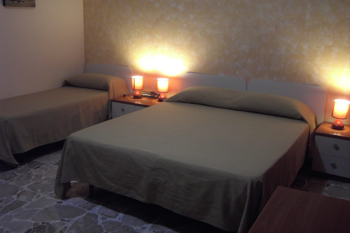A hotel room with two beds, each with a nightstand and a lamp. The room has a stone floor and a plain wall with a small picture.
