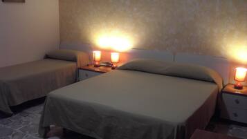 Habitación de hotel con dos camas, cada una con una mesita de noche y una lámpara. El piso es de piedra y la pared es sencilla, con un cuadro pequeño.